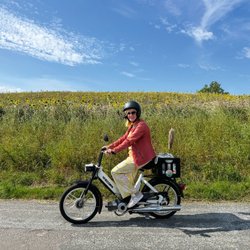 Eine Frau fährt auf eine Mofa an einer grünen Wiese vorbei.