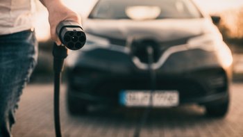 Person mit Ladestecker fürs Auto, im Hintergrund ein unscharfes Auto.