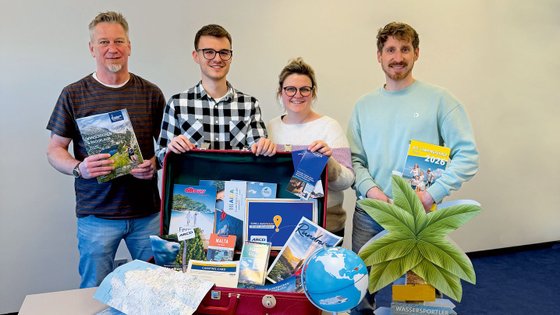 Eine Gruppe von vier Personen steht um einen roten Koffer mit vielen Reiseinformationen.