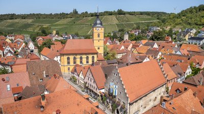 Blick aus der Vogelperspektive auf einen Ort mit historischen Häusern, Kirchturm und Weinbergen im Hintergrund