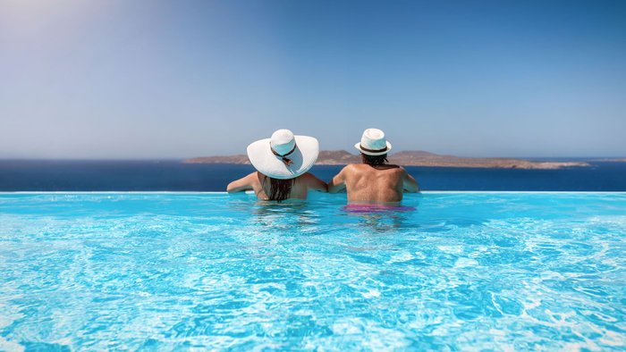 Paar mit Sonnenhüten genießt den Sommerurlaub in einem Infinity Pool mit Aussicht auf das Meer.