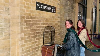 Zwei junge Frauen schieben scheinbar einen Gepäckwagen durch eine Backsteinwand unter einem Schild mit der Aufschrift „Patform 9 3/4