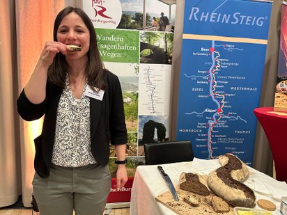 Frau vor Landschaftsbildern und Wanderkarte beißt in ein Butterbrot, neben ihr liegt ein Brotlaib in Schlangenform.