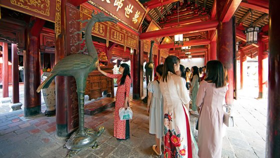 Asiatische Frau in einem in Rot und Gold gehaltenen Tempel streichelt einen Bronzekranich, um sie herum weitere Besucherinnen.