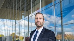 Ein Mann mit ernster Miene steht vor einem Gebäude mit Glasfassade.