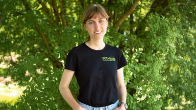 Junge Frau in T-Shirt und Jeans vor Baum mit grünen Blättern.
