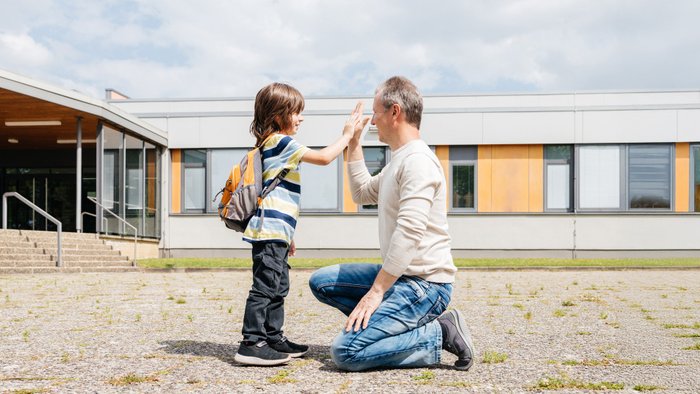 Ein Kind und sein Vater klatschen ab, im Hintergrund ein Schulgebäude