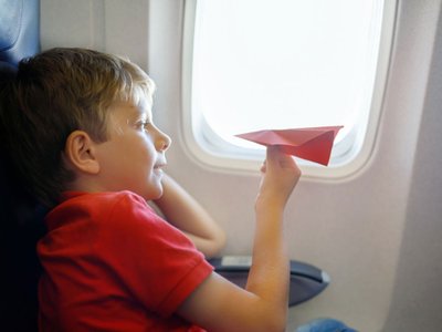 Kind sitzt im Flugzeug und spielt mit einem roten Papierflieger.