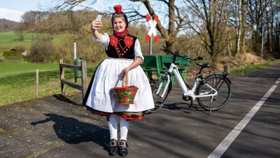 Mädchen in Rotkäppchen-Tracht steht am Radweg und macht ein Selfie.