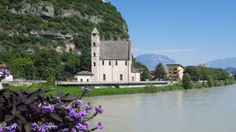 Blick auf den Fluss Etsch in Trento mit Blumen im Vordergrund und einer Kirche im Hintergrund.