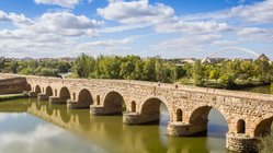 Landschaft mit römischer Steinbrücke über Fluss und moderner Stahlbrücke im Hintergrund.