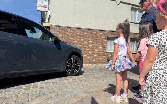 Kinder warten an der Straße, ein Auto biegt ab. 