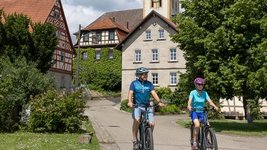 Zwei Radfahrer, im Hintergrund ein Dorf.