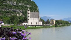 Blick auf den Fluss Etsch in Trento mit Blumen im Vordergrund und einer Kirche im Hintergrund.
