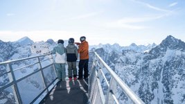 Drei Skifahrer stehen auf einem Aussichtssteg aus Stahl und genießen den Blick auf die verschneiten französischen Südalpen.