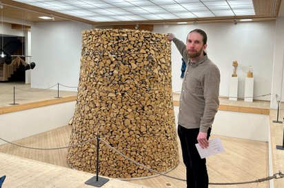 Mann zeigt in einem Museum eine zylinderförmige Installation, die aus vielen Holzscheiten besteht.