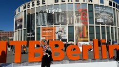 Frau steht vor ITB-Schriftzug, im Hintergrund Messeeingang mit bunten Werbeplakaten.