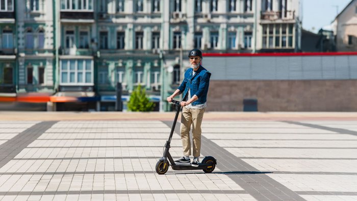 Mann mittleren Alters mit Sonnebrille und Helm fährt einen E-Scooter in einer Stadt.