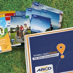 Ein ARCD-Urlaubspaket, daneben Reiseführer und Broschüren auf grünem Rasen.