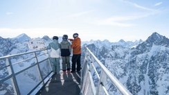 Drei Skifahrer stehen auf einem Aussichtssteg aus Stahl und genießen den Blick auf die verschneiten französischen Südalpen.