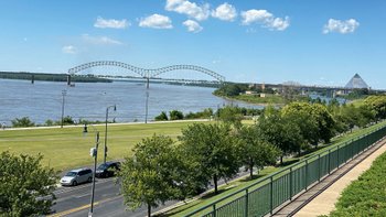  Blick auf Ufer des Mississippi mit Grünstreifen und M-förmiger Brücke im Hintergrund.