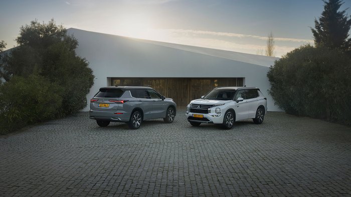 Ein grauer und ein weißer Mitsubishi Outlander stehen in der Abendsonne vor einem flachen Gebäude.