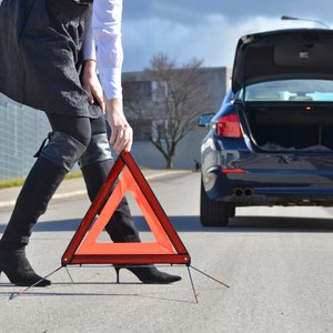 Mann stellt Warndreieck auf, im Hintergrund Auto mit offenem Kofferraum.