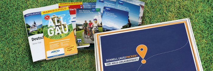 Ein ARCD-Urlaubspaket, daneben Reiseführer und Broschüren auf grünem Rasen.