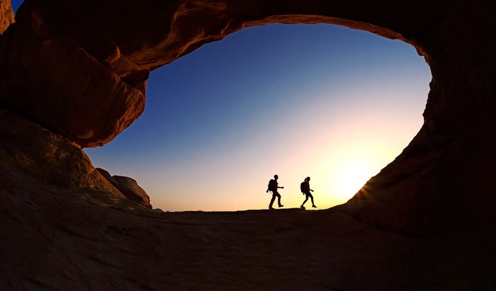 Zwei Wanderer – im Gegenlicht – laufen einen Felsbogen entlang.
