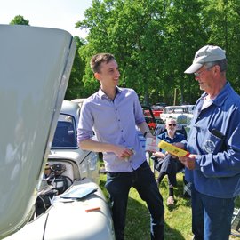 Zwei Männer, einer davon mit Flyer in der Hand neben einem Oldtimer mit offener Motorhaube.